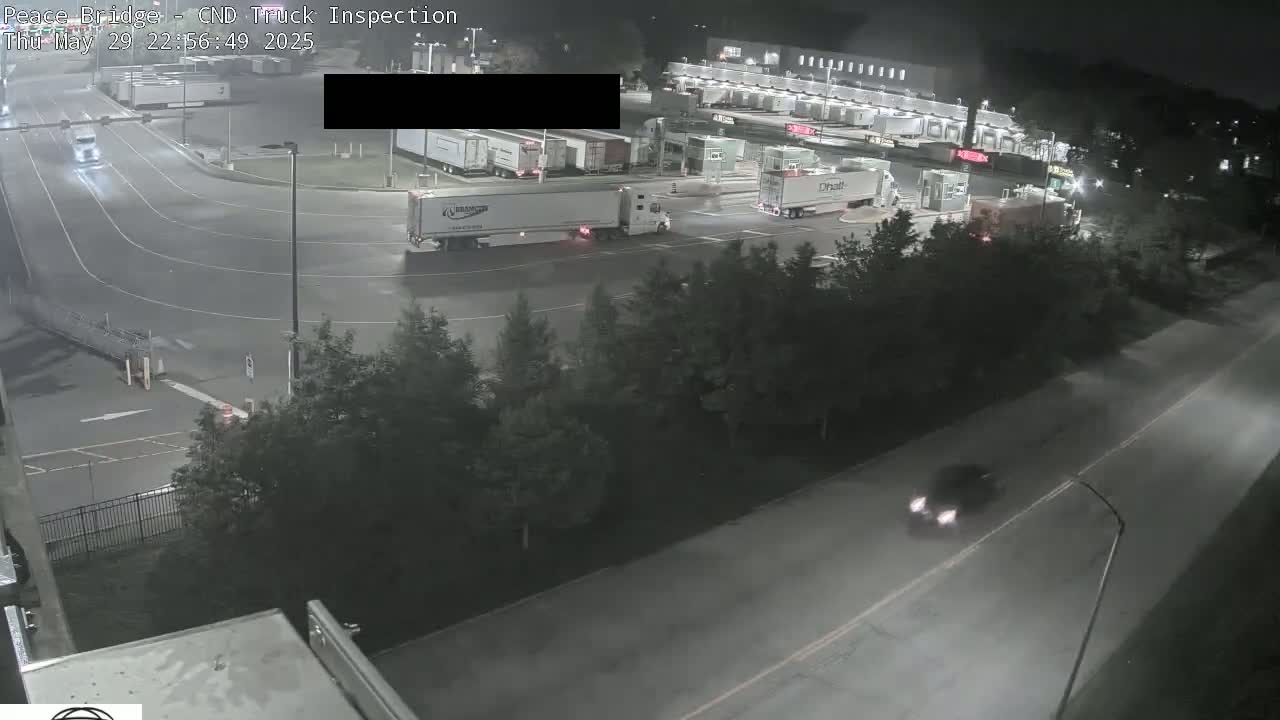 A nighttime aerial view shows several semi-trucks at a truck inspection station, with one car traveling on a nearby road.