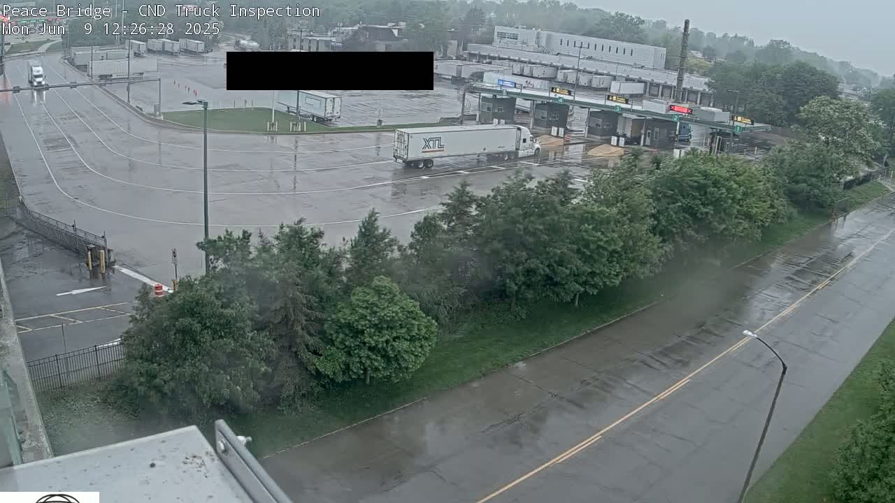 A wet, rainy view of a truck inspection station with several trucks parked and roadways, surrounded by green trees.