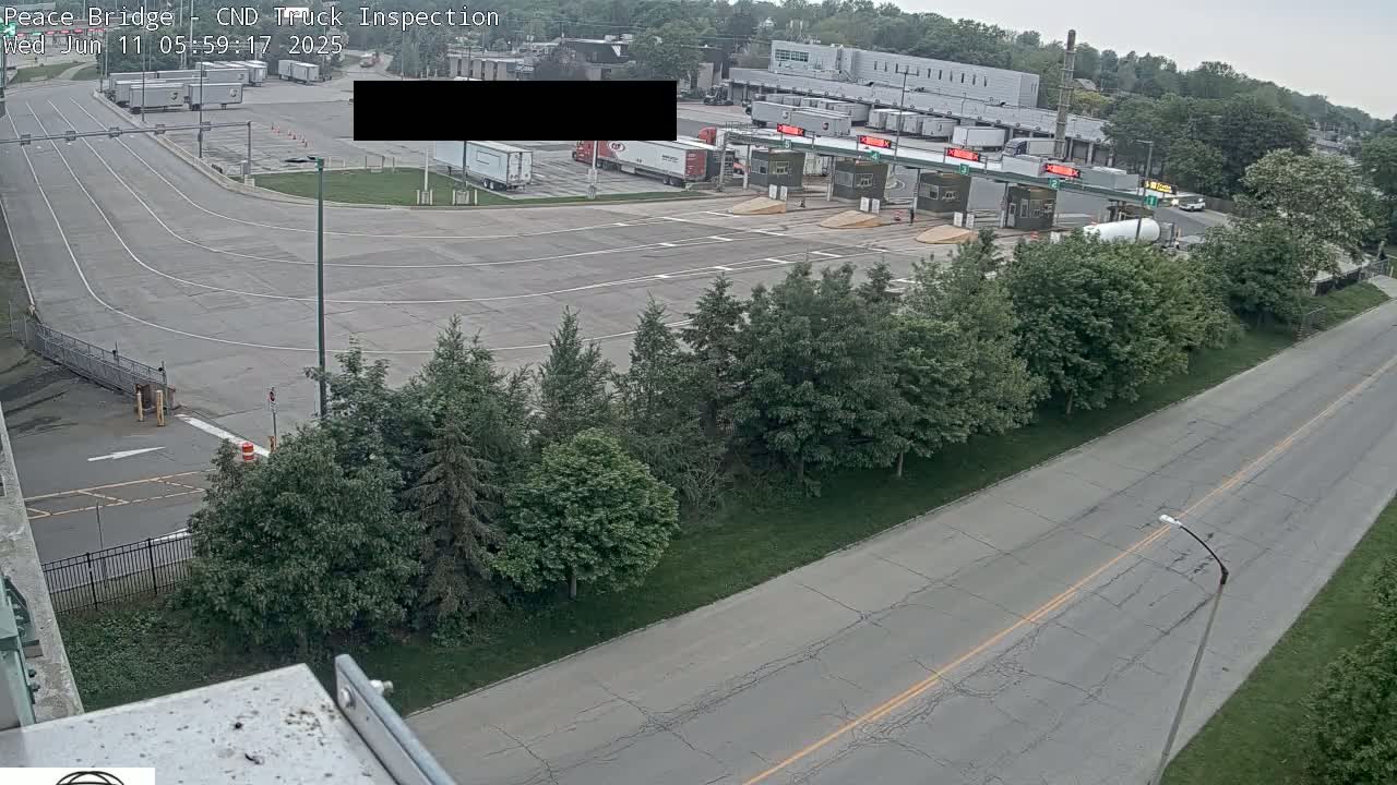 An aerial view shows a truck inspection station with several semi-trucks in a parking lot, a roadway leading to inspection booths, and surrounding green trees under an overcast sky.