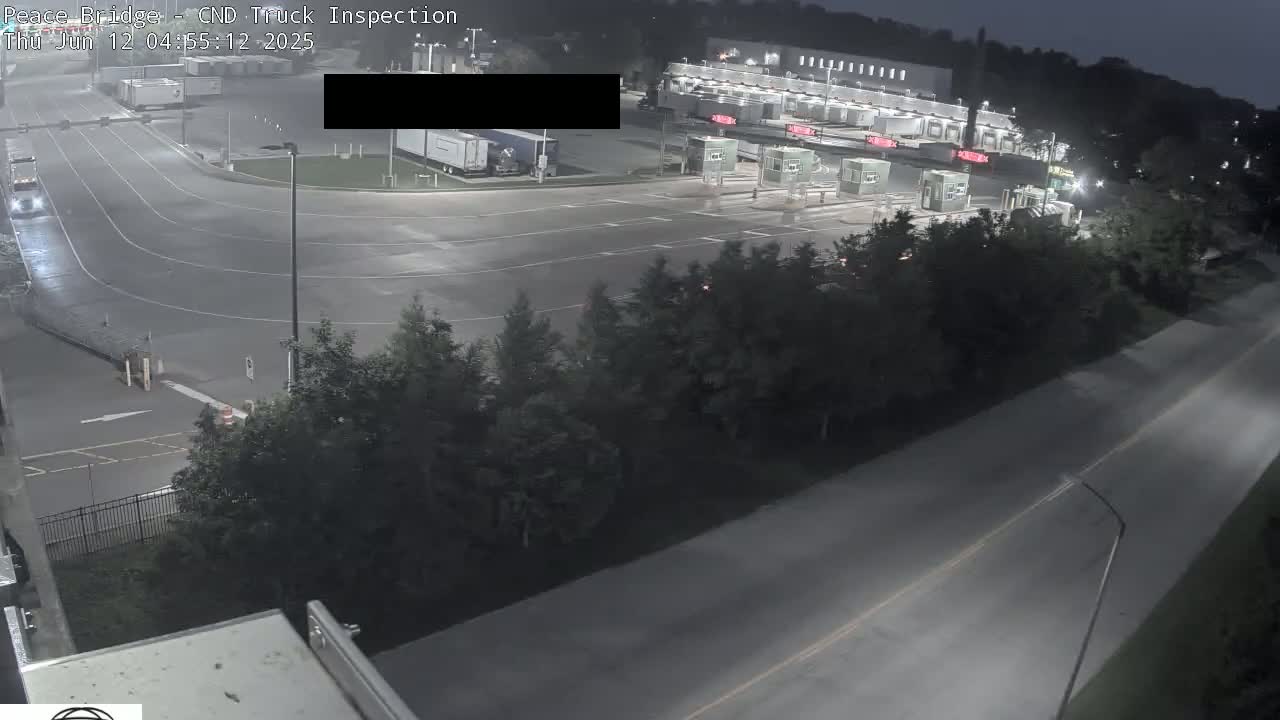 A nighttime aerial view shows a truck inspection station with several trucks and cars, surrounded by dark trees and a paved roadway.