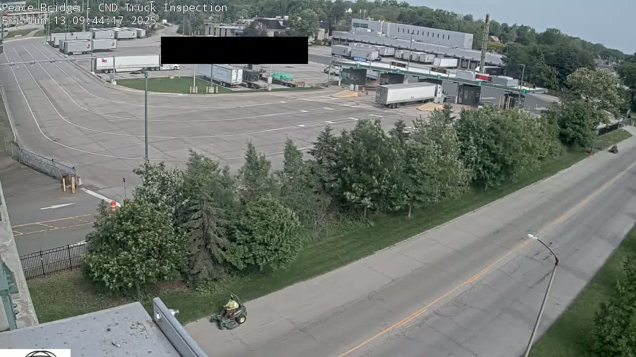 An aerial view shows a truck inspection station with numerous semi-trucks and a person operating a lawn mower on a sunny day.