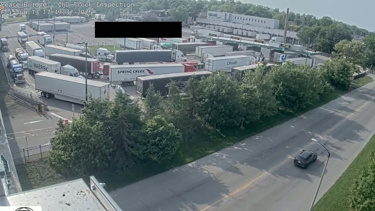A truck inspection area with numerous semi-trailer trucks parked and maneuvering, situated next to a road with one car visible, all under a sunny sky.