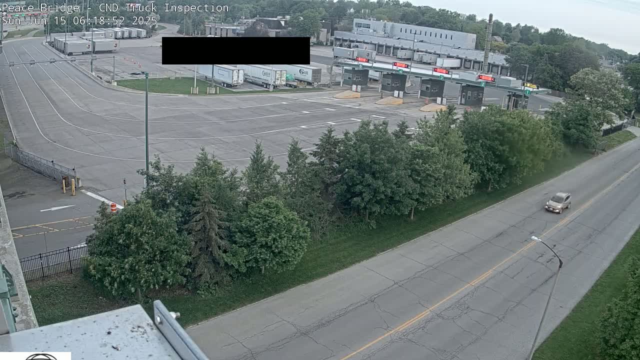 A high-angle view shows a truck inspection station with several semi-trucks parked nearby, a roadway with one car driving past, and green trees lining the sides of the road under an overcast sky.