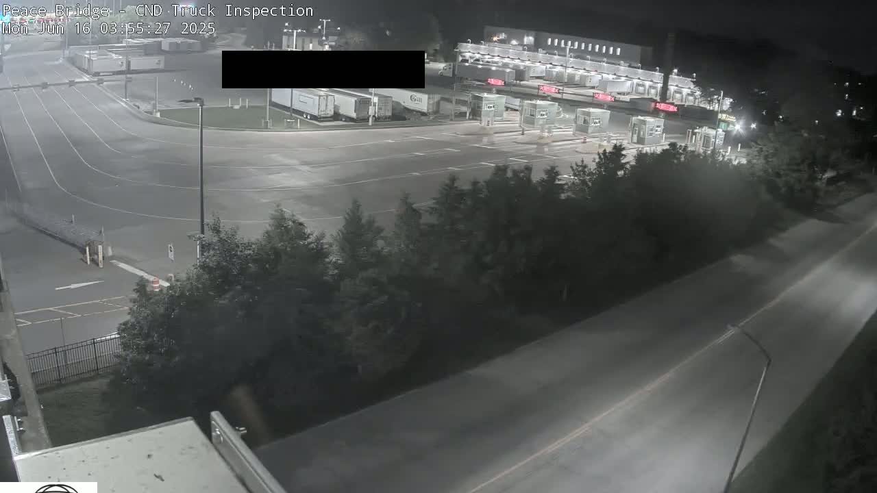 A nighttime aerial view shows a truck inspection station with several semi-trucks parked and a road leading to it, surrounded by trees.