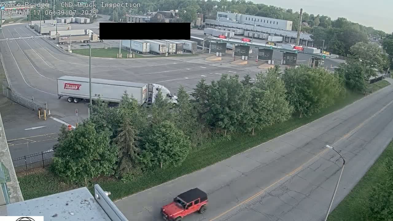 An aerial view shows a roadway with a red Jeep and several semi-trucks at a border crossing on a partly sunny day.