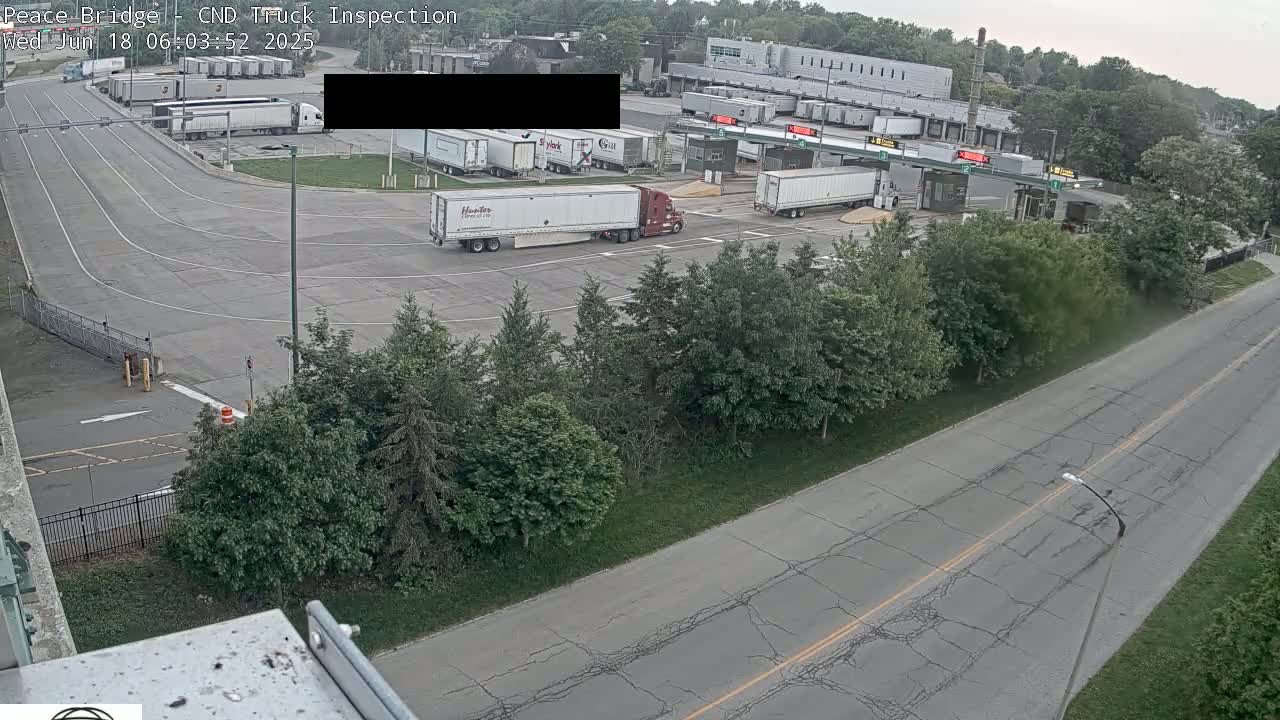 An aerial view shows a truck inspection area with numerous semi-trucks, some stationary and others in motion, under overcast skies.
