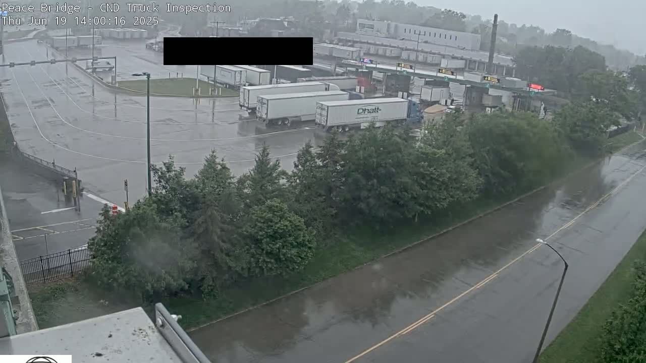 A rain-soaked truck inspection area with several semi-trailers parked, surrounded by green trees and a wet road.