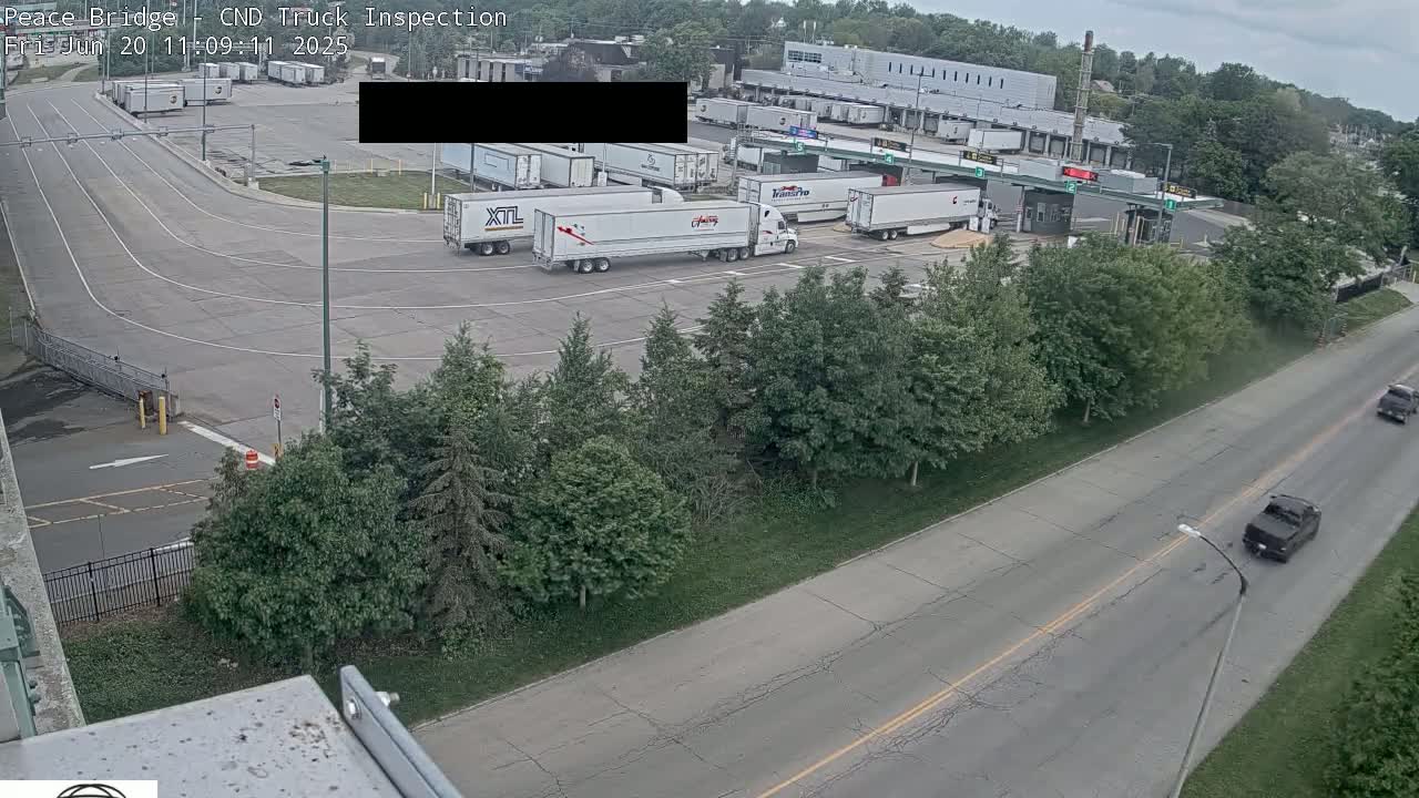 An aerial view shows several semi-trucks parked in a truck inspection area on an overcast day, with cars driving on a nearby road lined with trees.