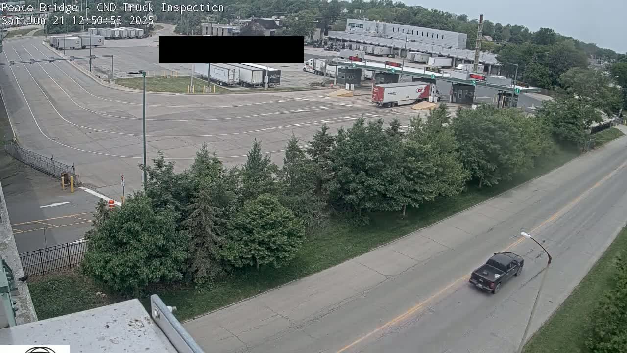 A high-angle view shows a truck inspection station with several semi-trucks parked in a large paved lot on an overcast day, with a single dark-colored pickup truck driving away on a nearby road.