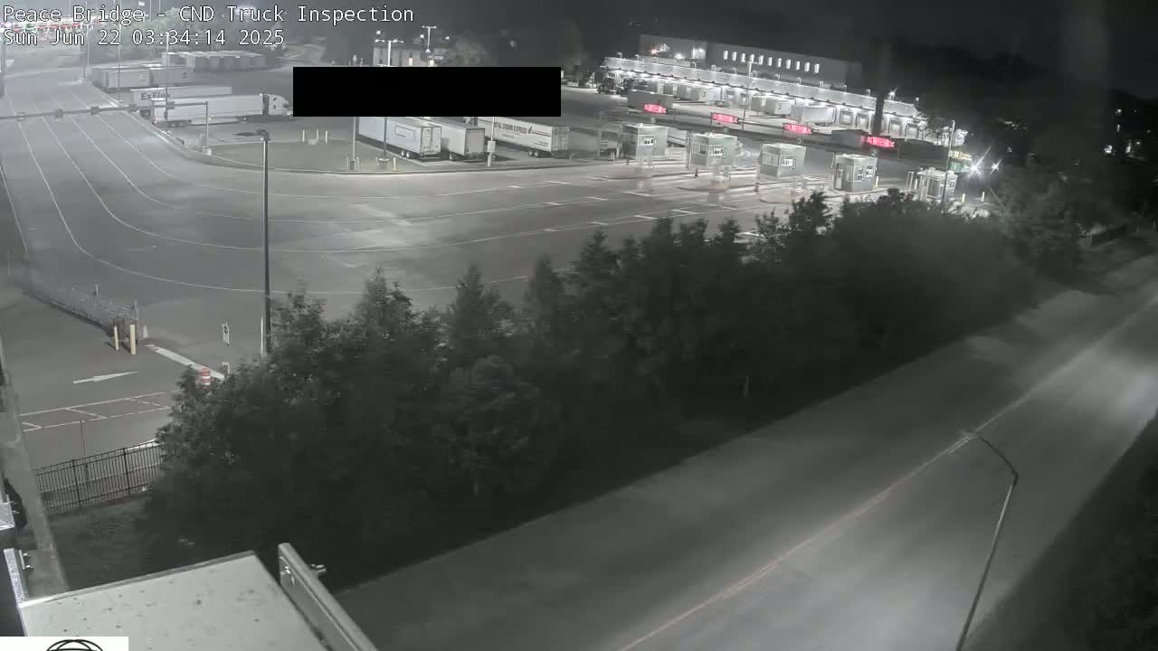 A nighttime aerial view of a truck inspection area at a border crossing, showing several semi-trucks parked and a roadway leading to inspection booths.