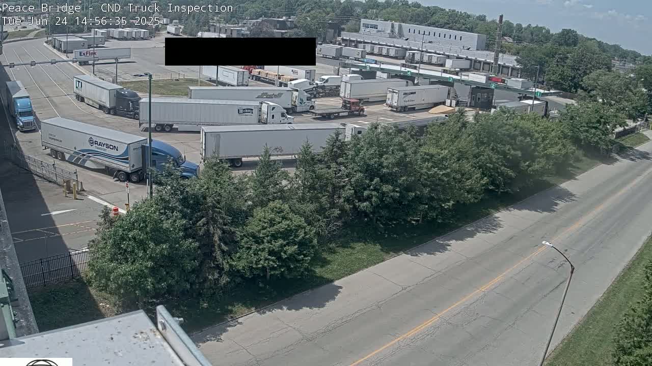 Numerous semi-trailer trucks are parked in a large lot on a sunny day, with a road visible in the foreground.