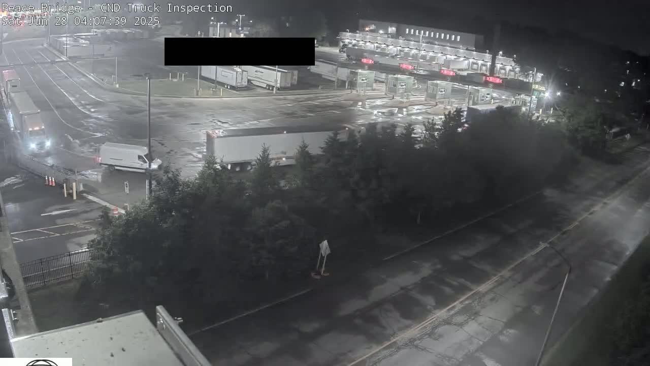A nighttime, wet scene shows a border crossing with several trucks and a van, surrounded by trees and roadway.