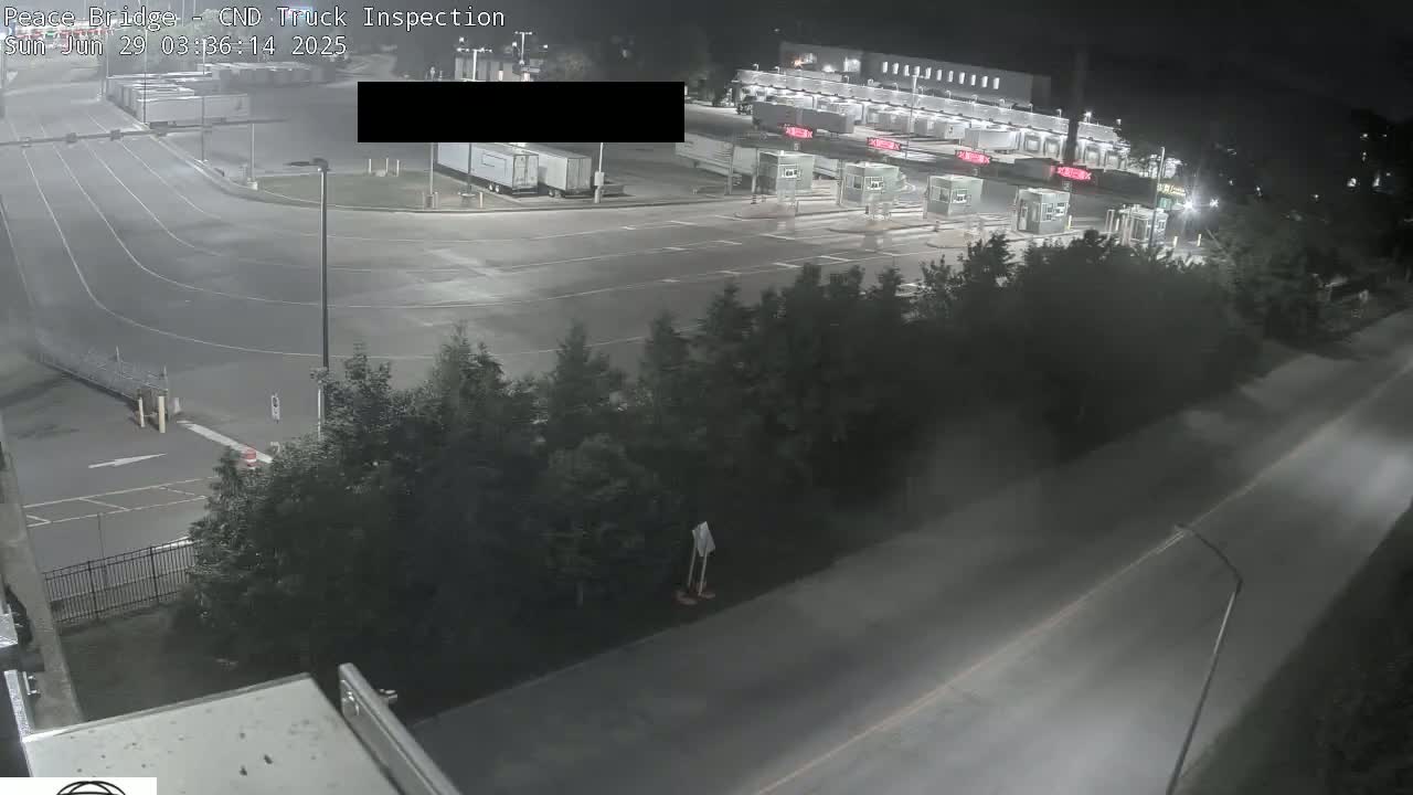 A nighttime overhead view shows a truck inspection station with several trucks and empty inspection lanes, surrounded by trees and roads under hazy conditions.