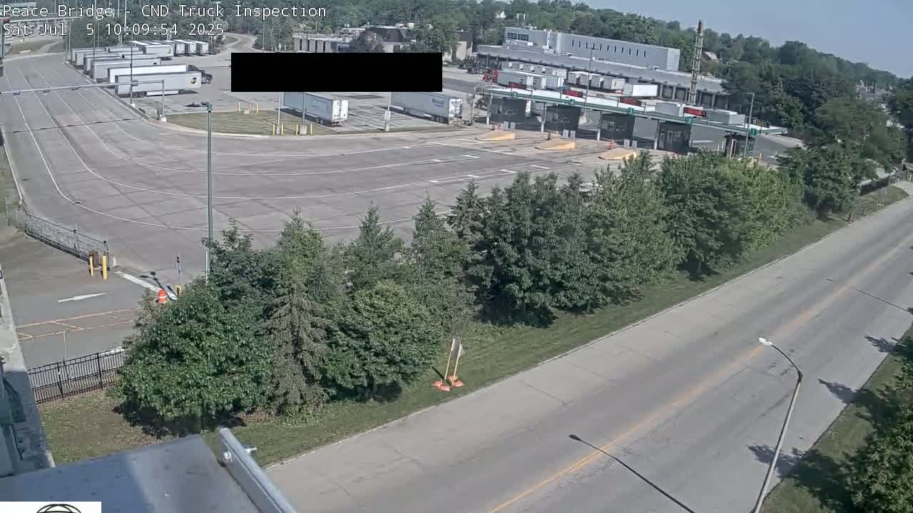 A truck inspection station with numerous semi-trucks, surrounded by trees and a roadway, under sunny skies.