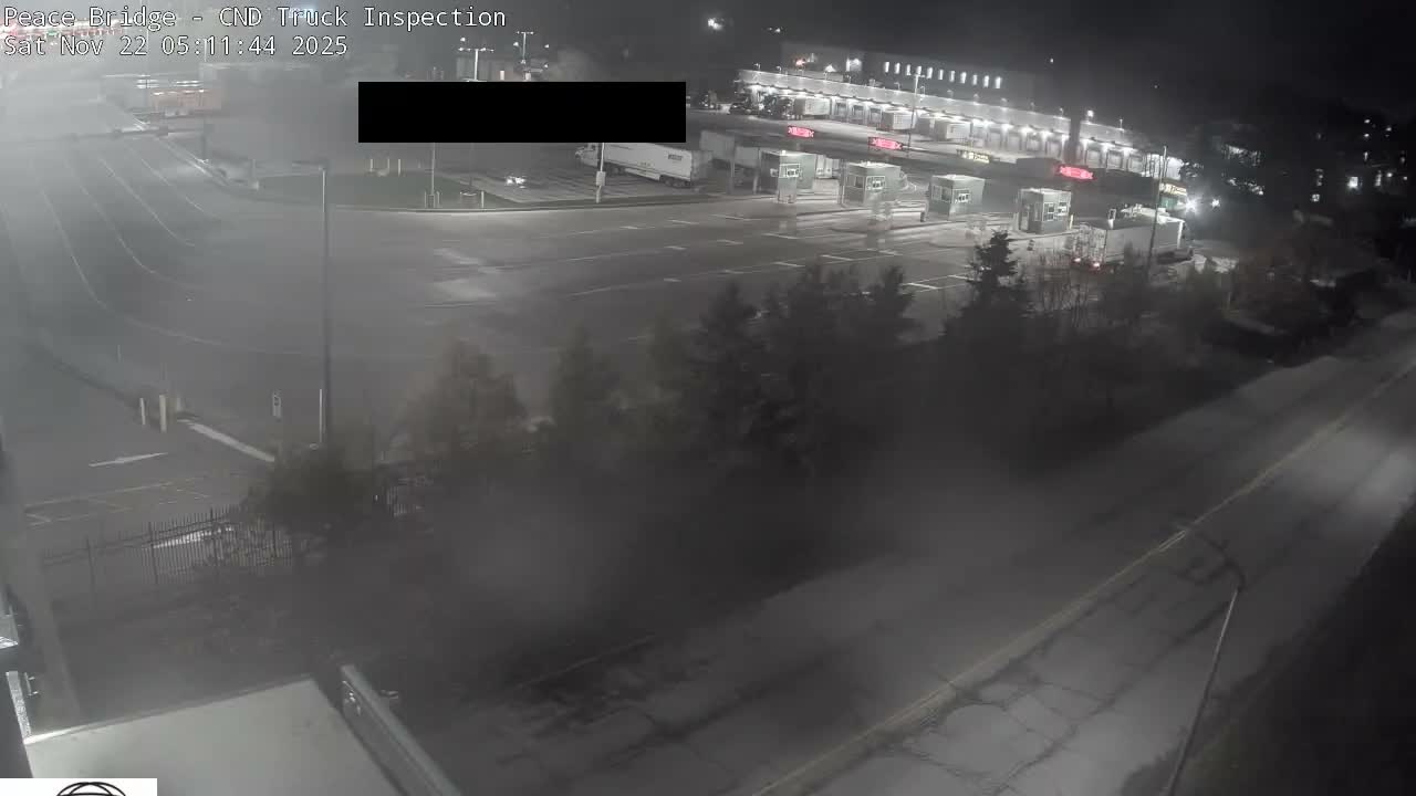 A dark, wet, and slightly hazy night scene depicts a multi-lane truck inspection facility with several booths, semi-trucks, and a brightly illuminated building in the background.