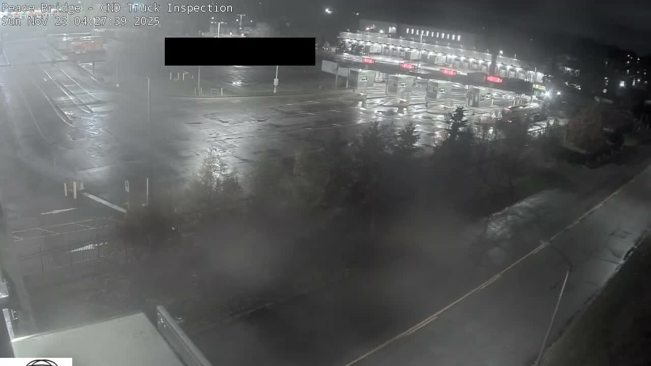 The image displays an overhead view of a well-lit vehicle inspection or border crossing facility at night, with wet asphalt and blurred distant details suggesting recent or ongoing rain.