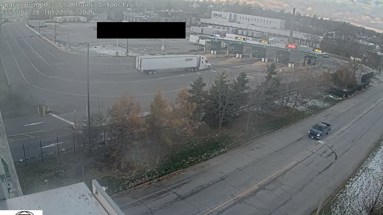 From an elevated perspective, a semi-truck approaches inspection booths at an outdoor facility, with a dark pickup truck driving on a cracked road nearby, under an overcast sky with patches of light snow or frost.