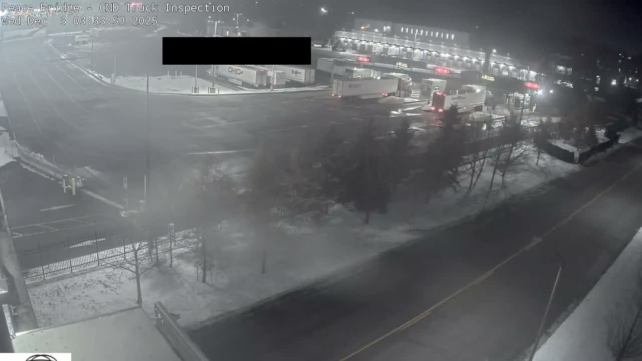 A snow-covered outdoor truck inspection facility is seen at night, with multiple semi-trucks parked and illuminated by artificial lights under hazy, wintry conditions.