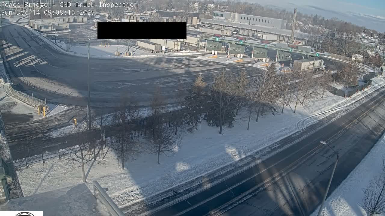 A snow-covered outdoor truck inspection facility is seen at night, with multiple semi-trucks parked and illuminated by artificial lights under hazy, wintry conditions.
