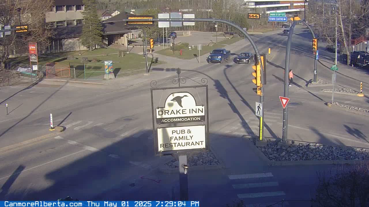 A sunny road intersection shows a Drake Inn sign, several cars, traffic lights, and pedestrians.