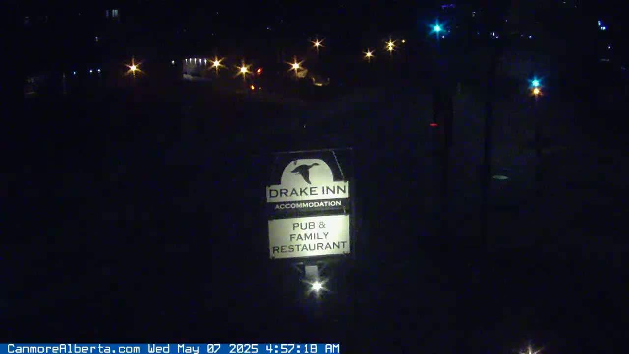 A dark nighttime scene shows a Drake Inn sign illuminated by a light source below it and distant streetlights in the background.