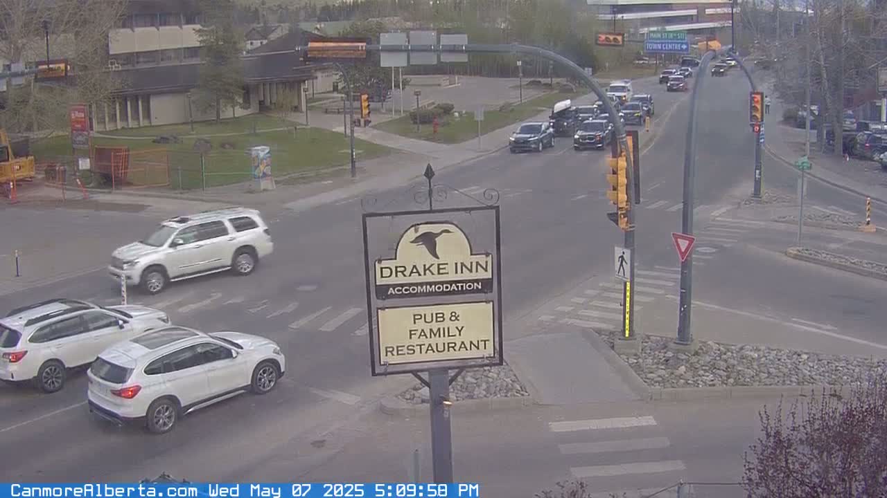 A partly cloudy day shows several vehicles at a crossroads near a pub and restaurant sign, with other buildings and trees in the background.