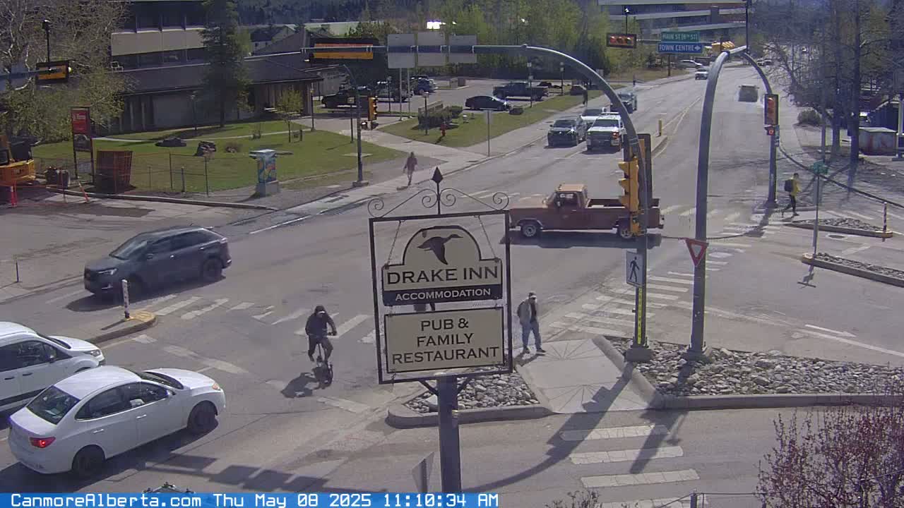 A sunny street scene shows a busy intersection with several vehicles, pedestrians, and a cyclist near a Drake Inn sign.