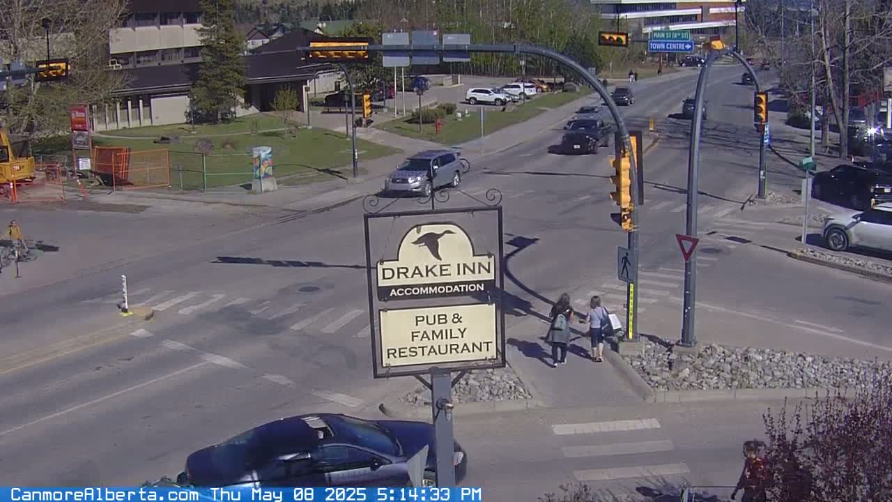 A sunny street scene shows a crossroads with several cars, pedestrians crossing a crosswalk near a Drake Inn sign, and a partly-constructed building in the background.