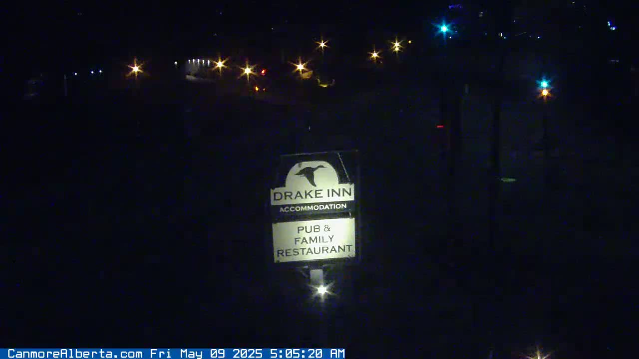 A nighttime view shows a Drake Inn sign illuminated by a light below it, with distant lights from a road visible in the background.