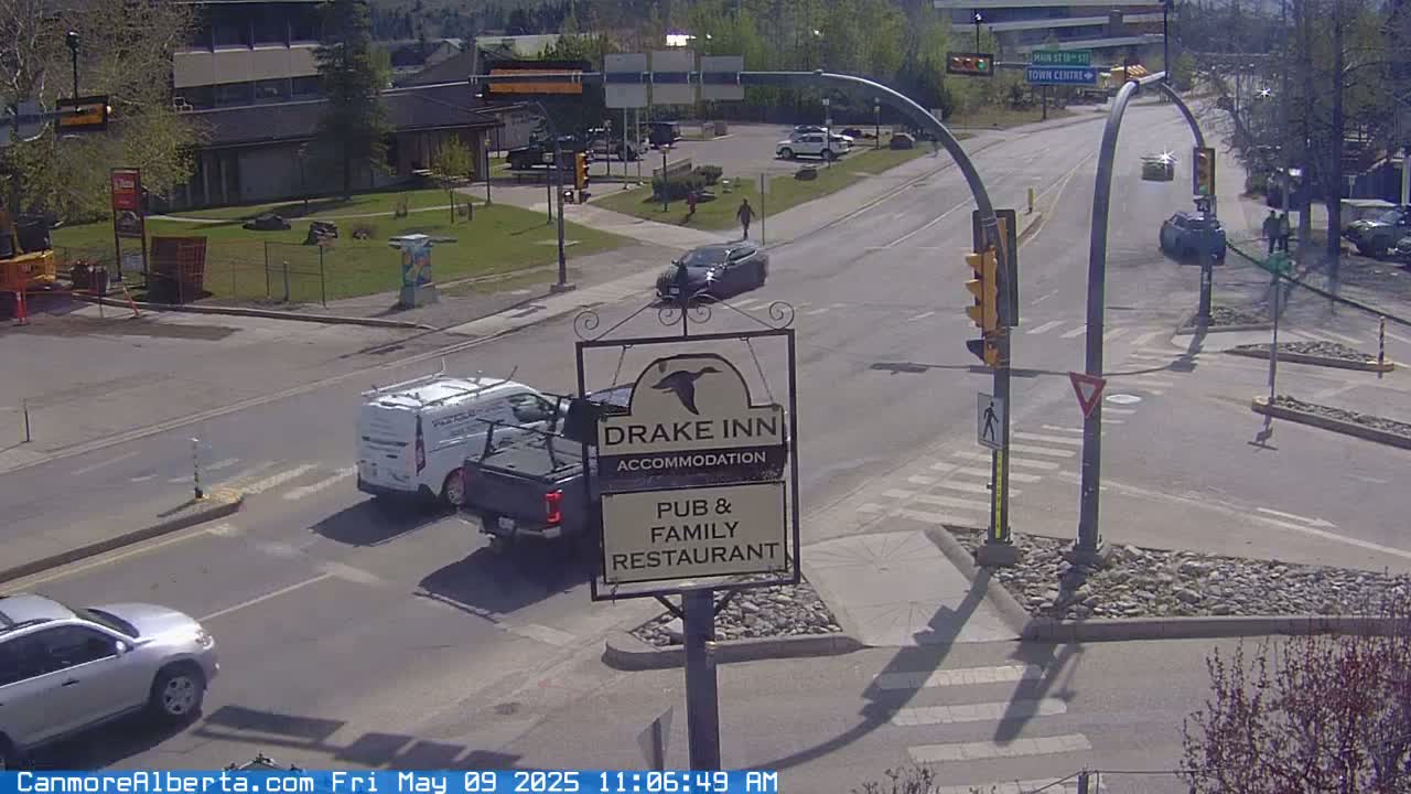 A sunny street intersection shows several vehicles, including a van and a silver SUV, with a pedestrian walking and a Drake Inn sign visible.