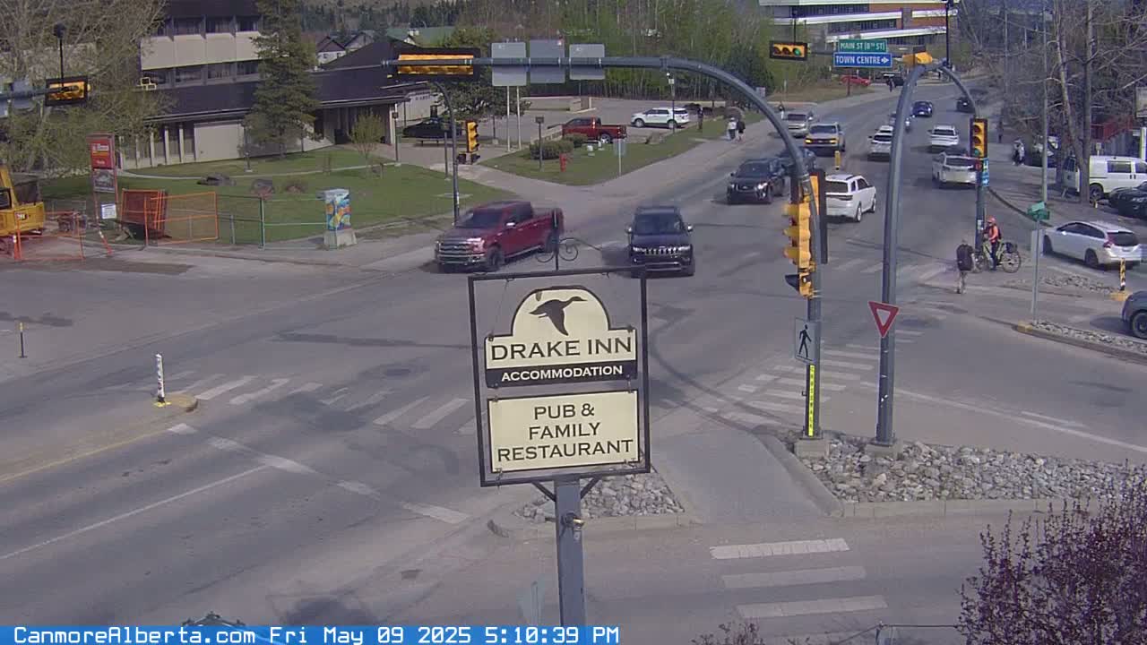 A sunny day shows a street intersection with several vehicles and pedestrians, including a person on a bicycle, near a Drake Inn sign.