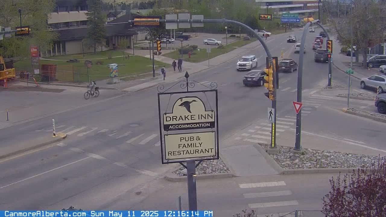 A sunny street scene shows a cyclist, pedestrians, and several cars at a multi-lane intersection with traffic signals and a Drake Inn sign.
