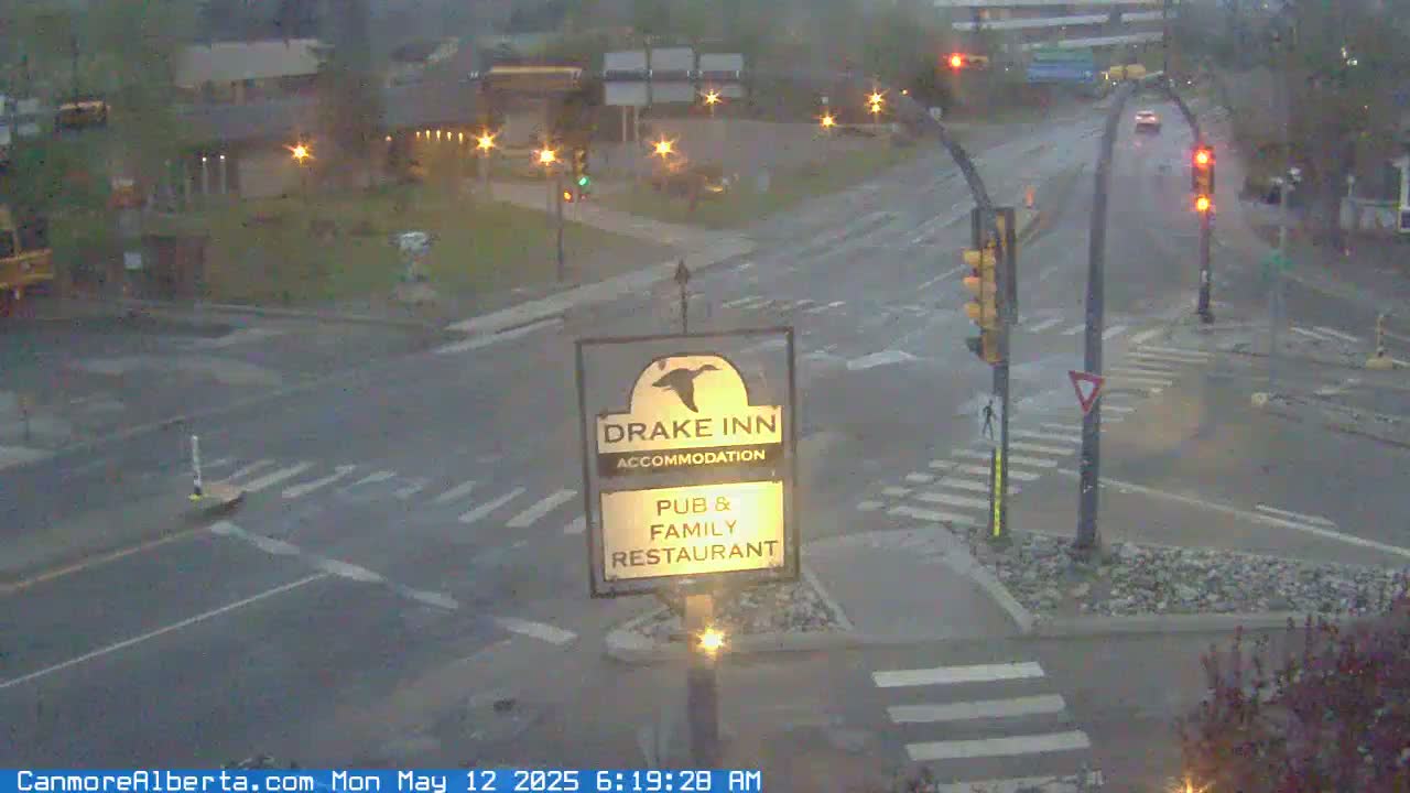 A wet road intersection with traffic lights and a Drake Inn sign in the foreground, under overcast skies.