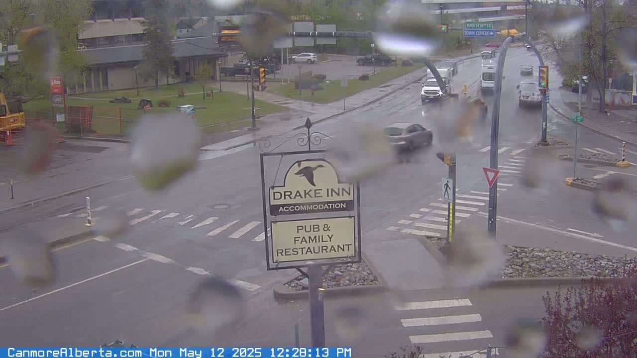 A rainy view of a street intersection with several vehicles, a pedestrian crossing, and a Drake Inn sign visible through rain-covered glass.