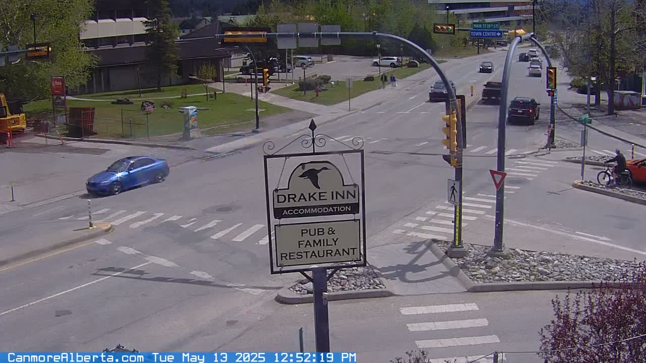 A sunny day shows a street intersection with a few cars and a person riding a bicycle, near a sign for the Drake Inn pub and restaurant.