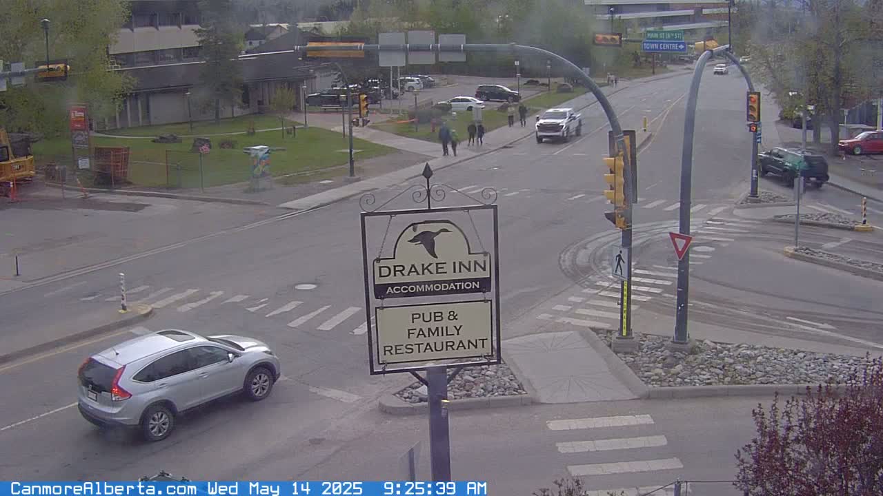 A silver SUV drives through a street intersection with a Drake Inn sign, pedestrians walking, and several other vehicles parked and driving, on an overcast day.