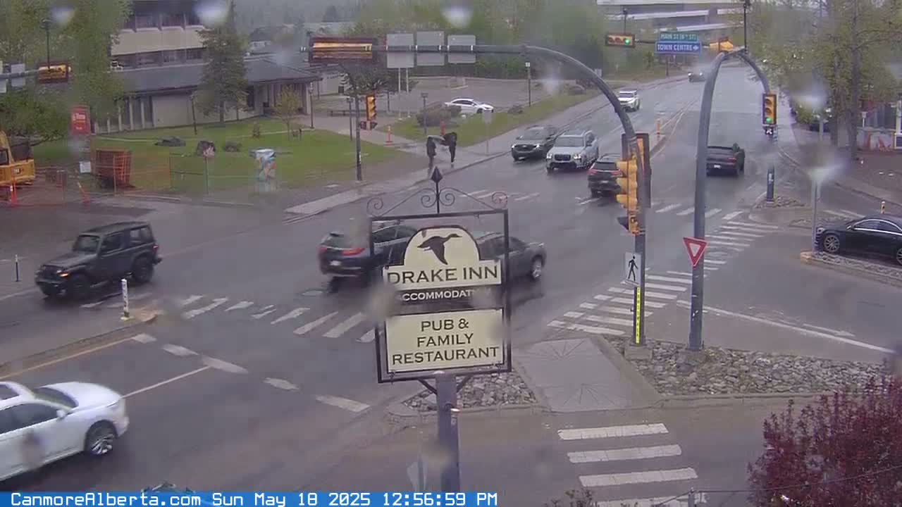 A rain-slicked intersection shows several vehicles and pedestrians near a Drake Inn pub and family restaurant sign.