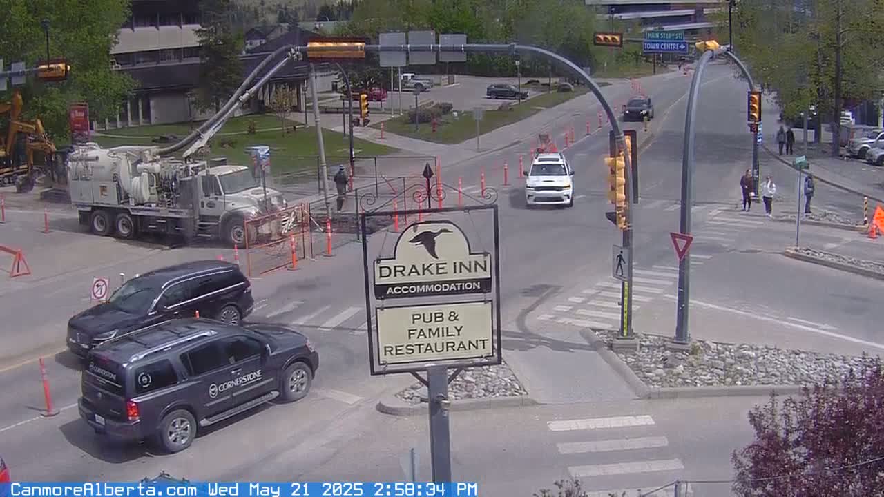 A street intersection with a road-cleaning truck, several vehicles, pedestrians, and a Drake Inn sign, on a sunny day.
