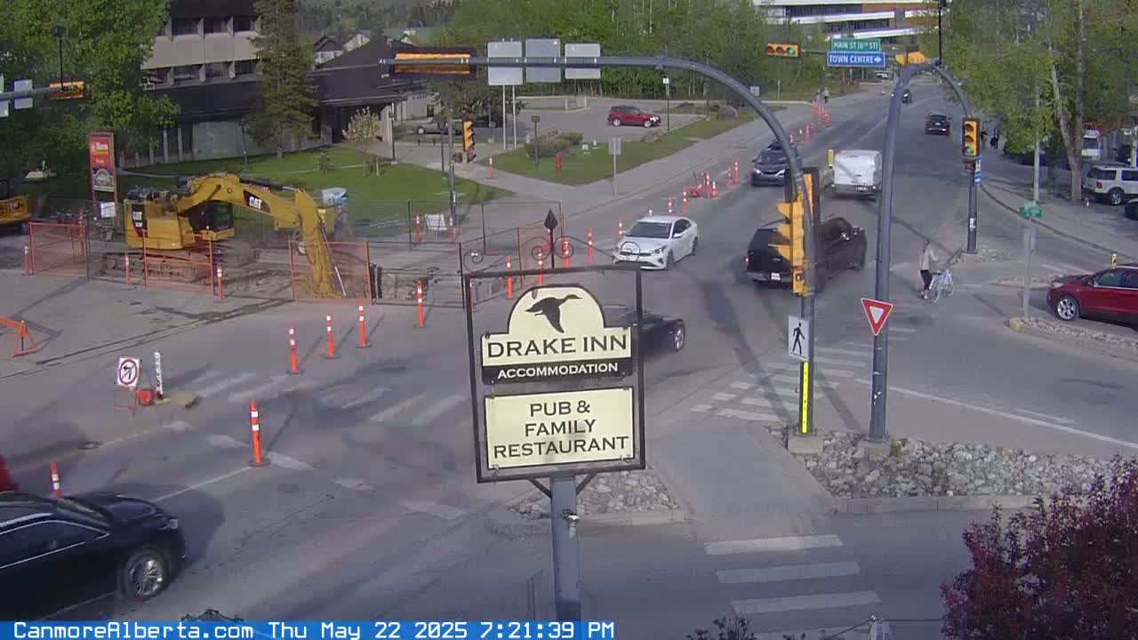 A sunny day shows a street intersection with several vehicles, a person riding a bicycle, construction equipment, and a Drake Inn sign.