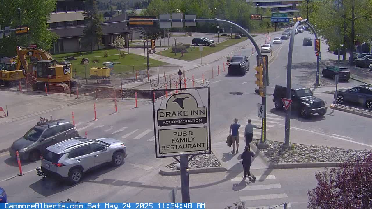 A sunny day shows construction equipment near a road intersection with several vehicles, pedestrians crossing, and a Drake Inn sign.