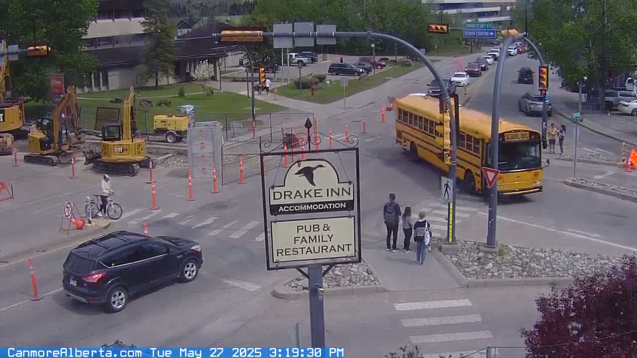 A sunny day shows a school bus stopped at an intersection with construction equipment nearby, several pedestrians walking, and a person riding a bicycle.