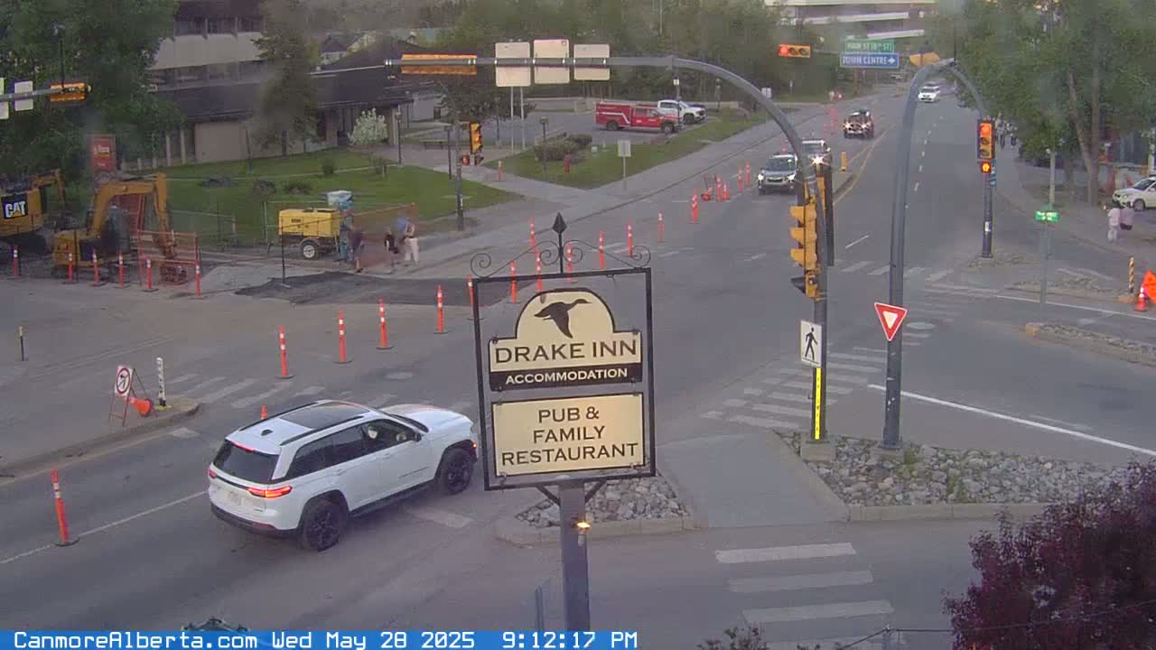 A street intersection with road construction, a white SUV, and several cars driving under partly cloudy evening conditions.