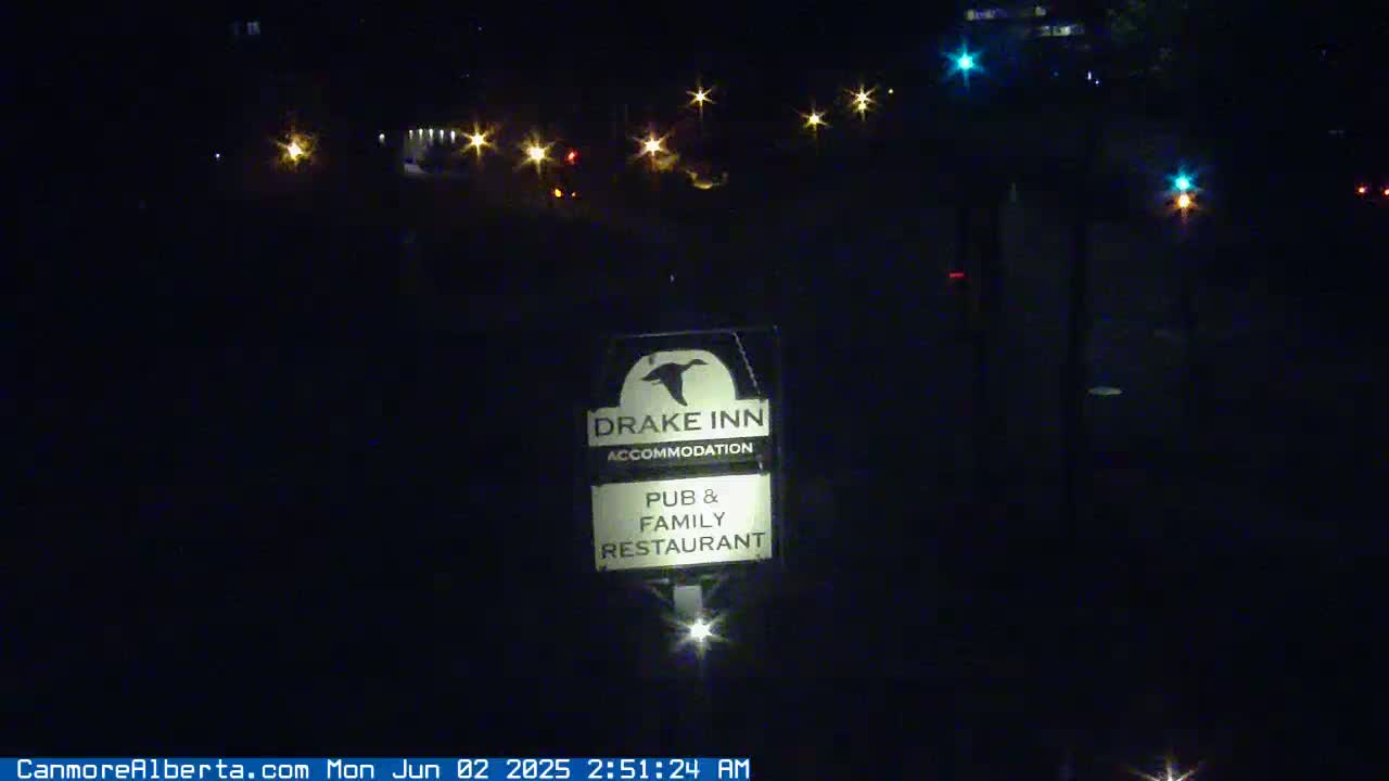 A nighttime image shows a Drake Inn sign illuminated by a light below it, with distant car lights visible in the background under a dark sky.
