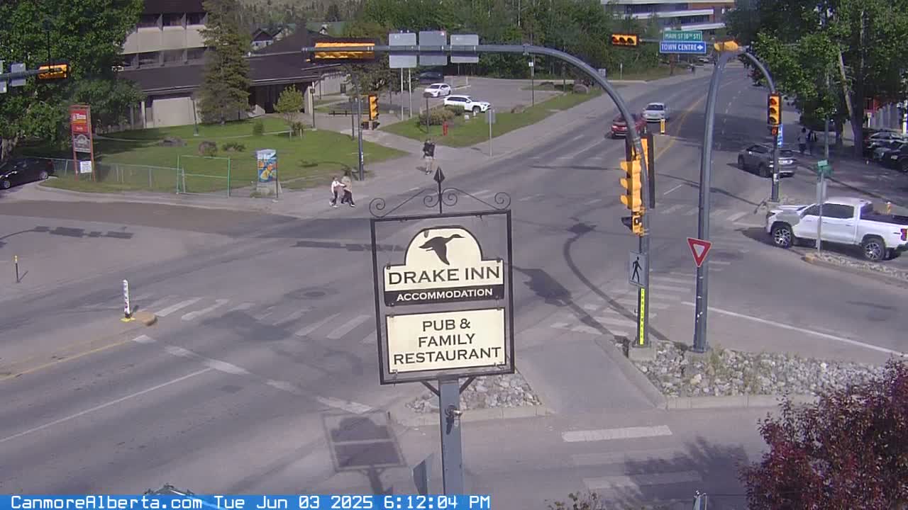 A sunny day shows a street intersection with several vehicles and pedestrians, near a sign for the Drake Inn pub and family restaurant.