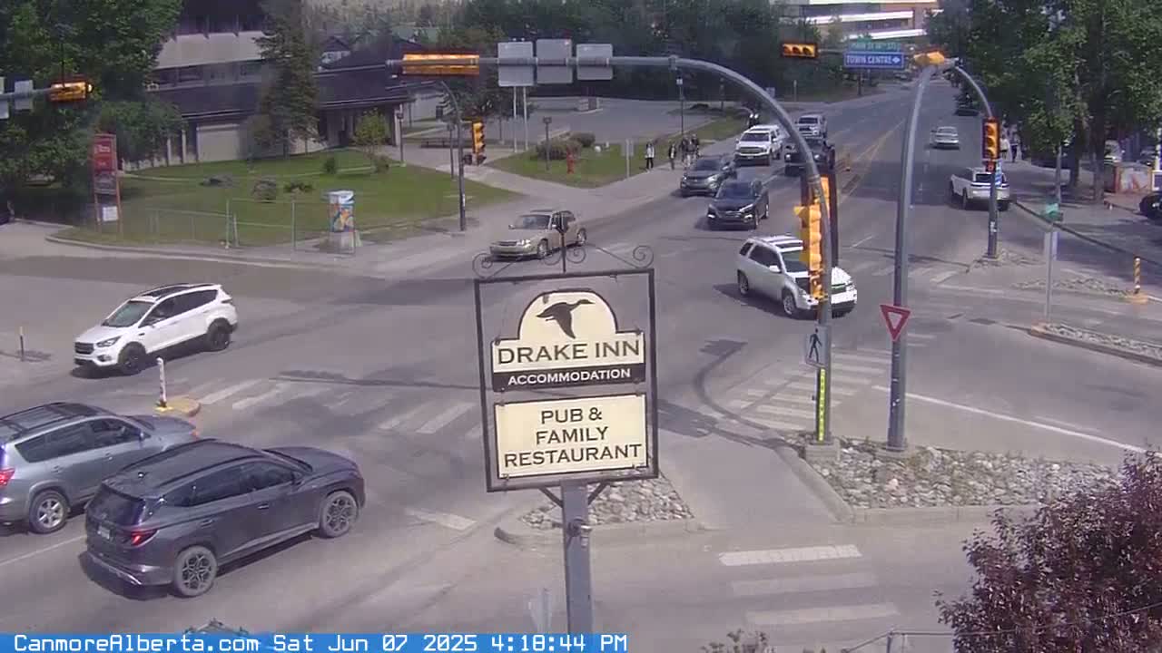 A street intersection with several cars, a Drake Inn pub and family restaurant sign, and a pedestrian crossing, on a sunny day.