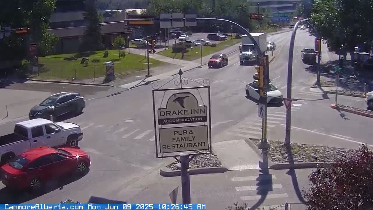 A sunny day shows a road intersection with several cars, a pickup truck, and a Drake Inn sign advertising accommodation, pub, and family restaurant.