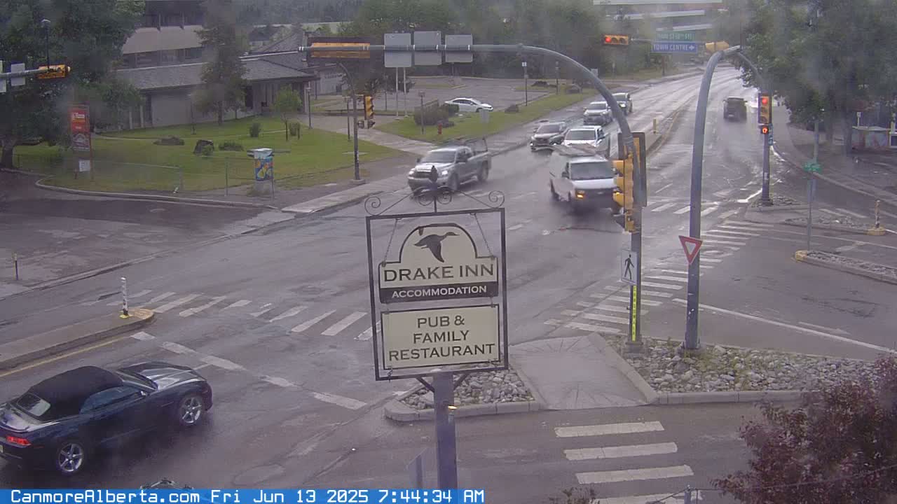 A wet, overcast street intersection with several vehicles, including a dark convertible, and a Drake Inn sign.