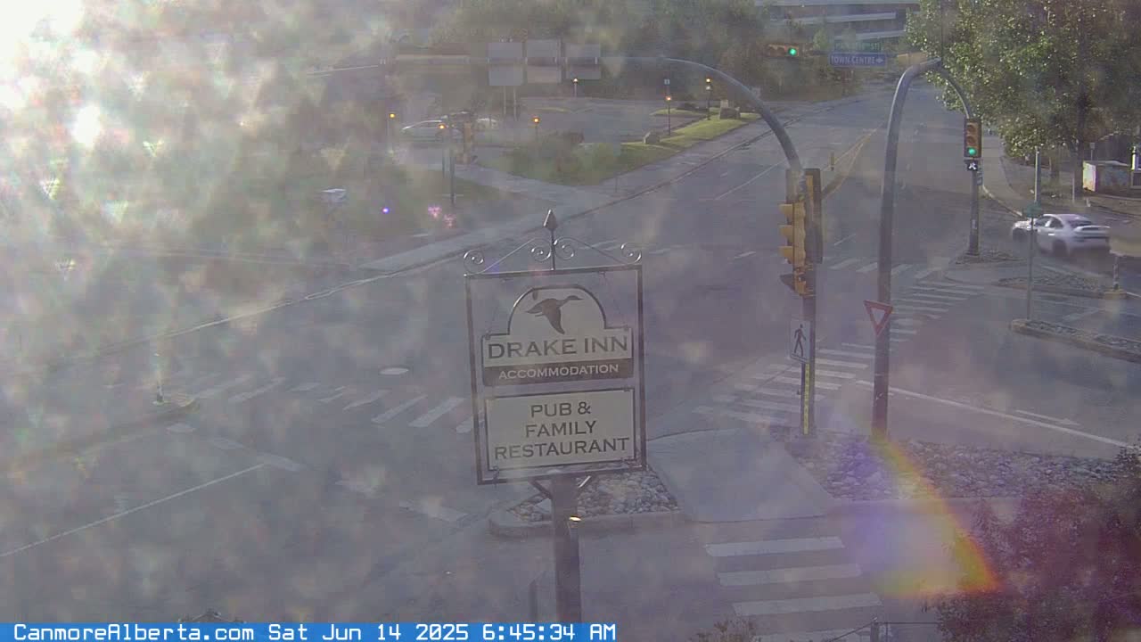 A hazy, sunlit street scene shows a Drake Inn sign, a car driving by, and a crosswalk with traffic signals.