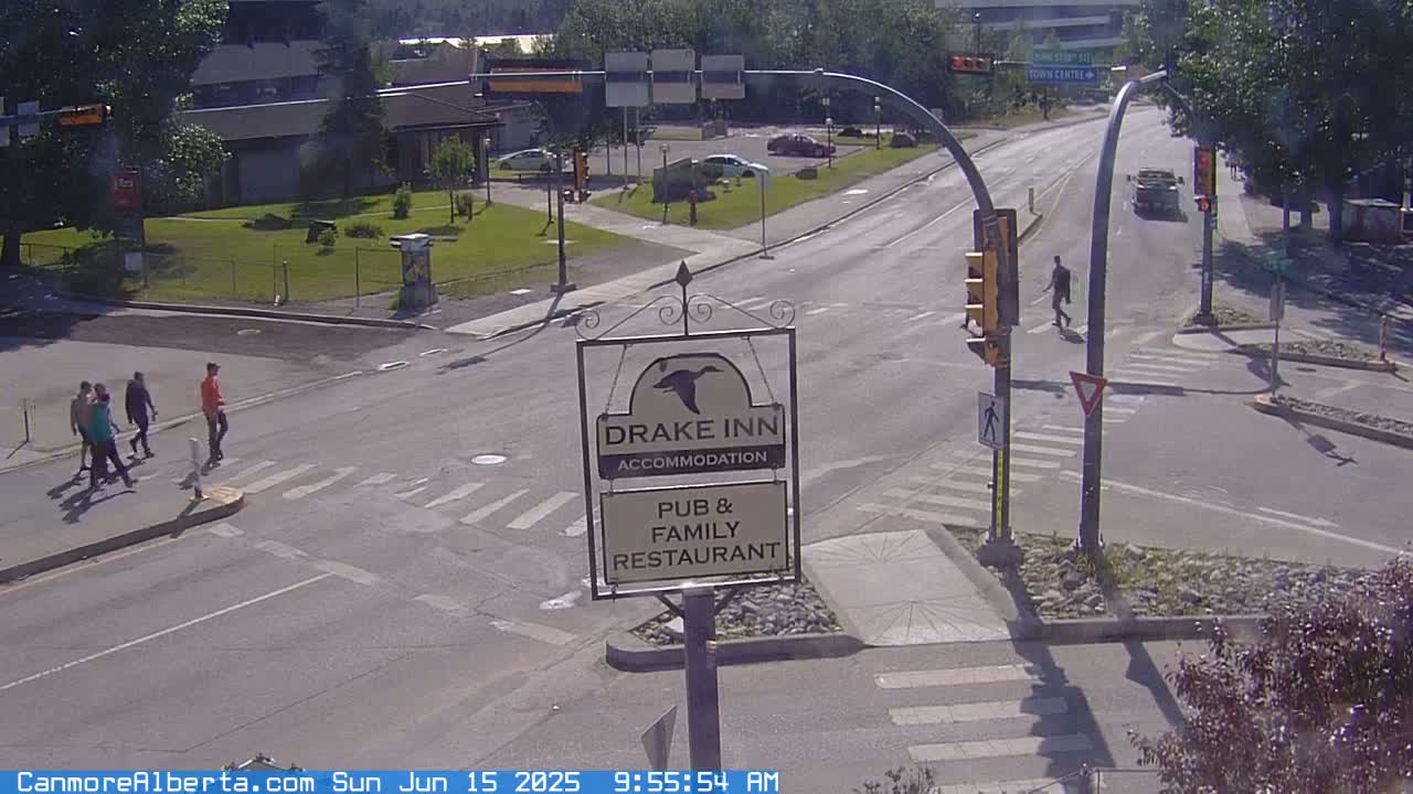 A sunny street intersection shows a Drake Inn sign, pedestrians crossing, and several cars.