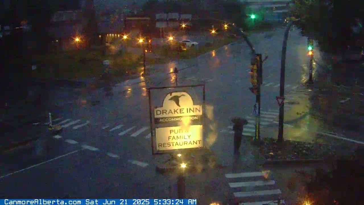 A rain-soaked street intersection at night, showing a Drake Inn sign and a pedestrian crossing.